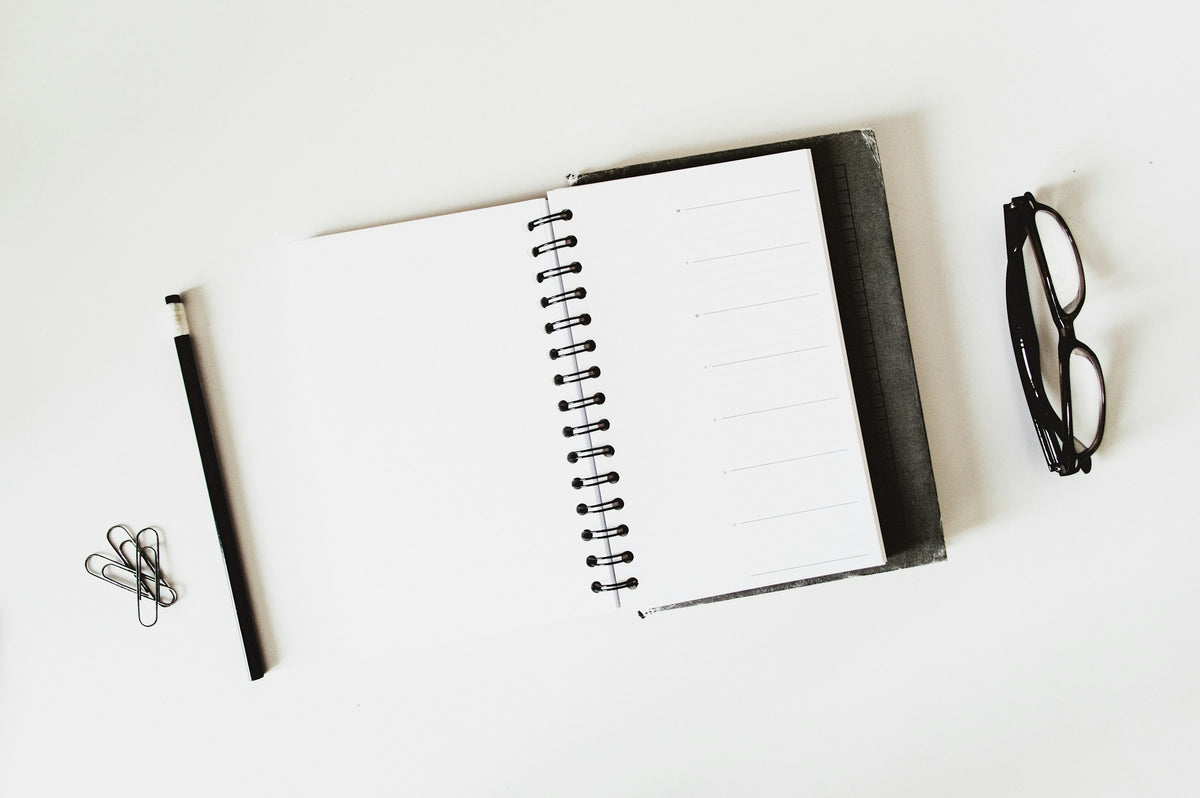Open spiral notebook with a black pencil, paper clips and eyeglasses on a white desk.