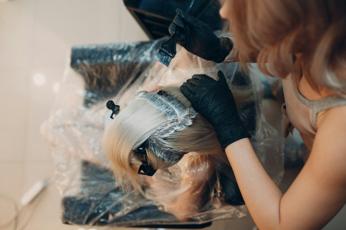 Overhead view of a gloved stylist applying color to sectioned hair with a tint brush.